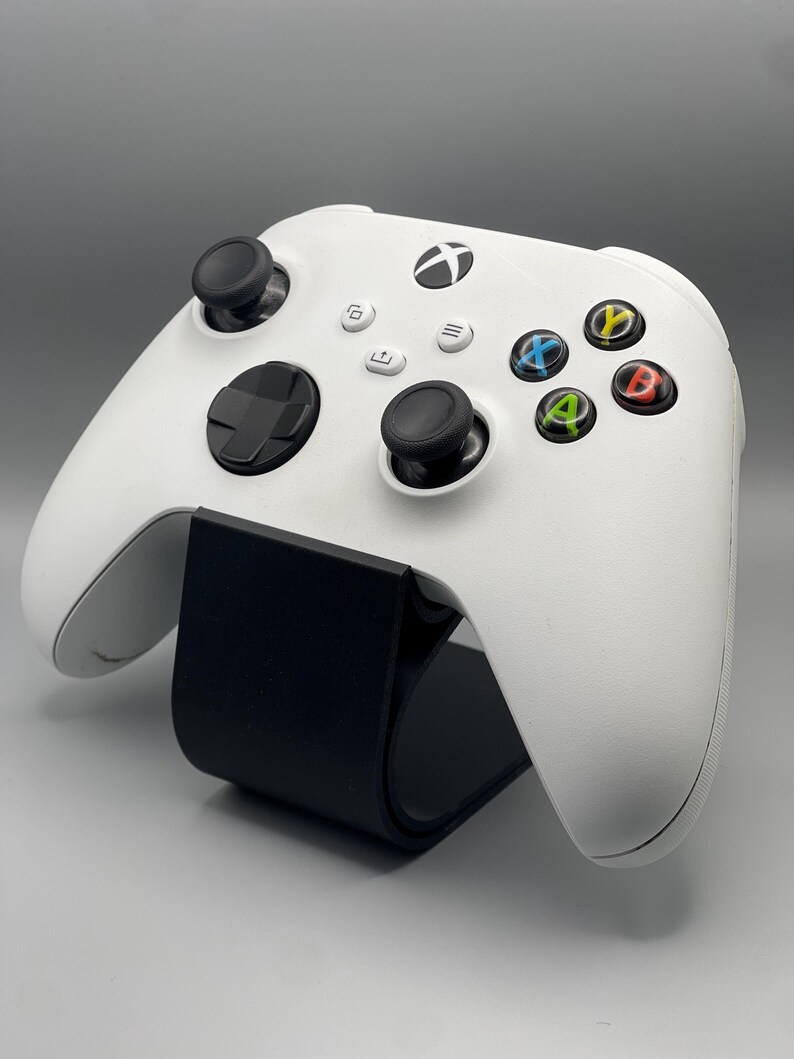 May include: A white Xbox Series X controller with black buttons and a black stand. The controller has a black Xbox logo on the front.