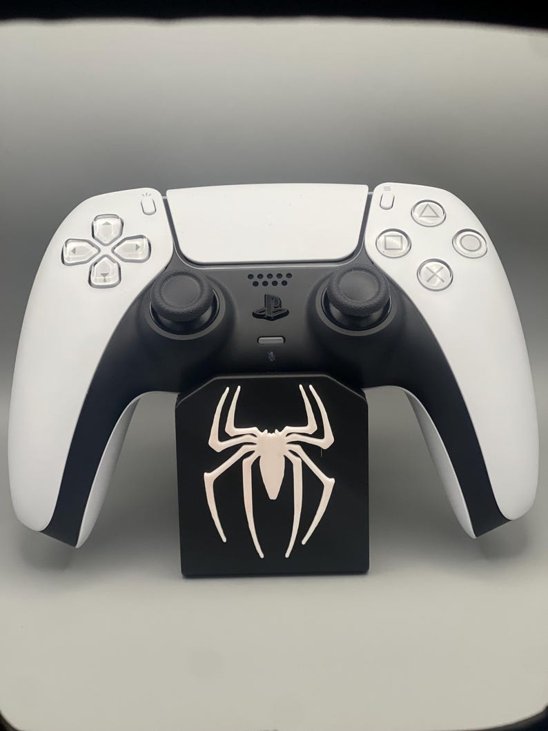 May include: A white PlayStation 5 DualSense controller with black accents is resting on a black stand with a white spider logo.