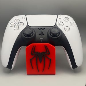 May include: A white PlayStation 5 controller with black accents is resting on a red stand with a black spider logo.