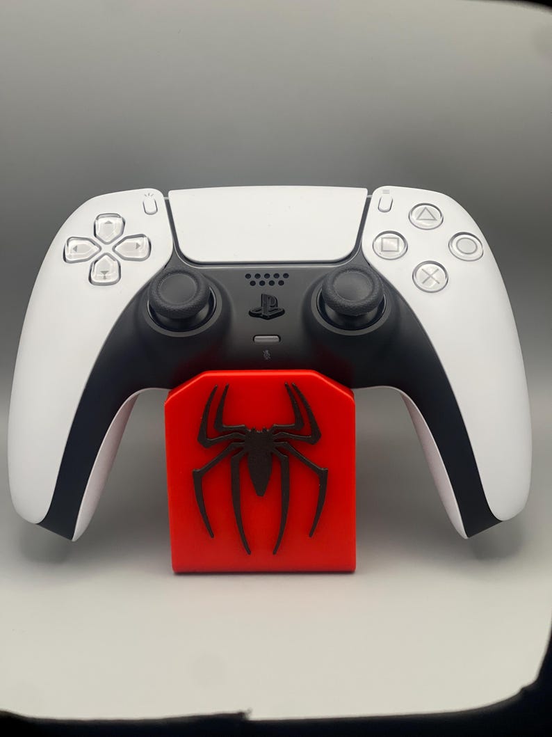 May include: A white PlayStation 5 controller with black accents is resting on a red stand with a black spider logo.