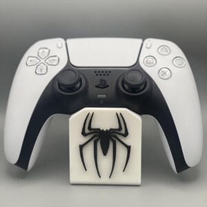 May include: A white PlayStation 5 controller with a black spider logo on a white stand.