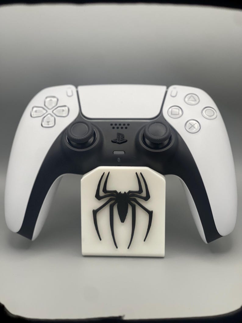 May include: A white PlayStation 5 controller with a black spider logo on a white stand.