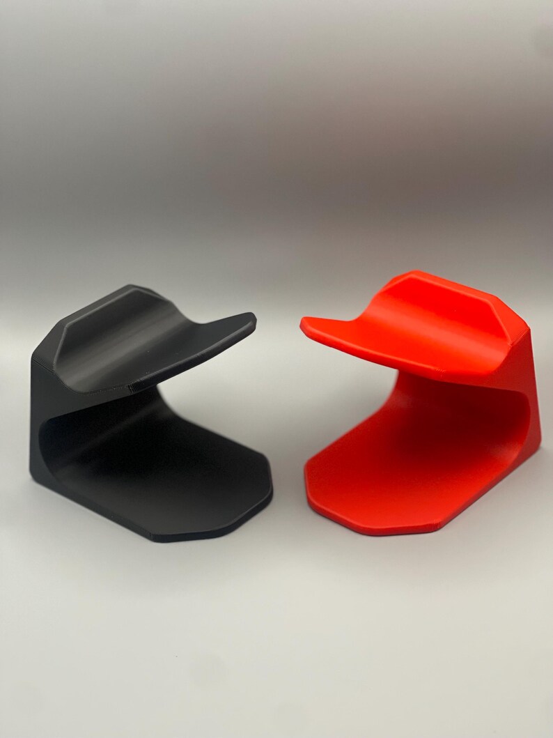 May include: Two 3D printed phone stands, one black and one red. The stands are designed to hold a phone upright.
