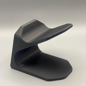 May include: A black plastic phone stand with a curved design. The stand is designed to hold a phone upright.
