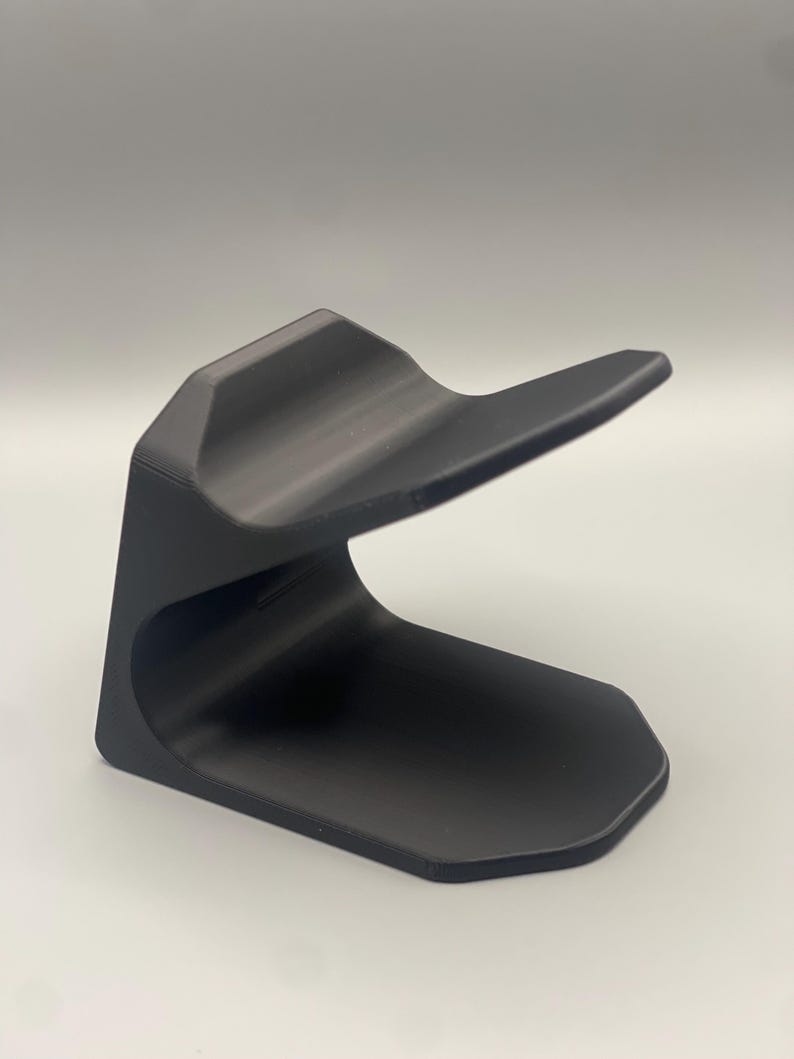 May include: A black plastic phone stand with a curved design. The stand is designed to hold a phone upright.
