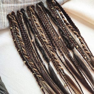May include: A collection of synthetic hair extensions featuring a mix of dark brown, light brown, and blonde strands. The extensions are styled into various braids and twists, accented with small metallic gold beads throughout the lengths.