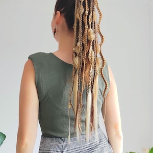 Può includere: Una donna con lunghe trecce bionde con accenti marroni e biondi. Le trecce sono legate insieme con un elastico per capelli marrone e hanno piccole perline dorate e ciondoli marroni lungo tutta la lunghezza. La donna indossa una canotta verde e una gonna a quadri neri e bianchi.