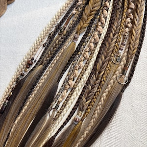 Puede incluir: Primer plano de múltiples extensiones de cabello trenzado en varios tonos de marrón, beige y rubio. Algunas trenzas están adornadas con cuentas y anillos metálicos. Las trenzas están dispuestas en diagonal sobre un fondo neutro.