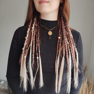 May include: Braided hair extensions in shades of brown and blonde, adorned with wooden beads. A matching necklace with a circular pendant and a black long-sleeved top complete the look. The hair extensions are styled in multiple braids.