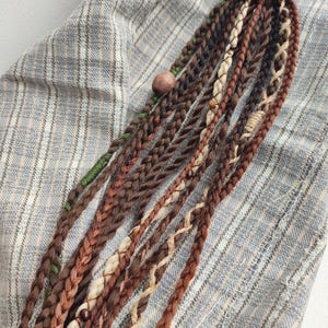 Puede incluir: Un conjunto de extensiones de cabello marrón, blanco y verde con cuentas y una banda elástica marrón. Las extensiones están dispuestas sobre un fondo de tela a rayas.