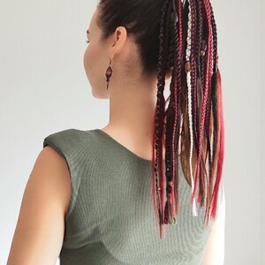 May include: A person with long, dark brown hair with red, white, and black braids tied up in a ponytail. The braids are decorated with wooden beads.
