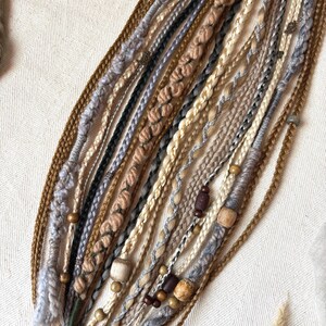 May include: A collection of handmade dreadlocks in various shades of brown, beige, gray, and black. The dreadlocks are adorned with wooden beads and decorative wraps, showcasing a bohemian style. Braided and twisted textures create a unique, textured appearance.