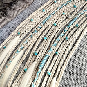 Op de afbeelding: Een verzameling blonde haarverlengingen met turquoise kralen en witte en blauwe borduursels. De verlengingen zijn gerangschikt in rijen op een grijze stoffen achtergrond. De tekst "27HairTreasures" is zichtbaar in de rechterbovenhoek van de afbeelding.