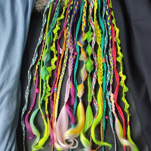 May include: A selection of brightly coloured hair extensions, featuring braids in shades of pink, yellow, green, orange, and blue. The extensions are arranged to cascade downwards, displaying different braiding techniques and decorative details. The overall look is vibrant and playful.