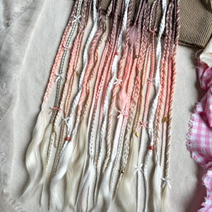 Puede incluir: Colección de extensiones de cabello trenzadas en tonos blanco, rosa y marrón. Cada trenza está adornada con pequeños lazos blancos, cuentas y abalorios. Los extremos de las trenzas son ondulados y de color claro.