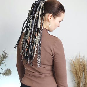 May include: A woman with dark hair wearing a brown turtleneck, with a ponytail of dreadlocks. The dreadlocks are a mix of black, gray, and cream colors, adorned with wooden beads and decorative elements. The background is a plain white wall.