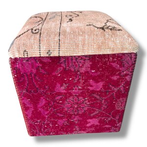 May include: A square ottoman upholstered in two distinct sections of woven textile. The top surface features a light beige fabric with dark brown linear and floral patterns, while the base is covered in a vibrant magenta fabric with intricate floral motifs.