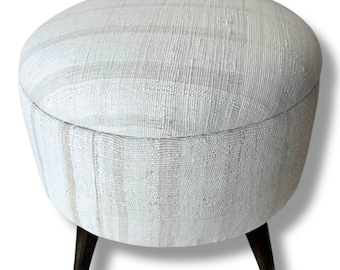 May include: A round ottoman upholstered in a textured, off-white fabric with subtle vertical stripes. The piece features a flat, circular top and is supported by three dark brown, tapered wooden legs that extend outward from the base.