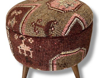 May include: A round ottoman upholstered in a textured, woven fabric featuring a multi-colored geometric pattern in shades of brown, dark red, and soft pink. The piece is supported by three short, tapered wooden legs with a natural brown finish.