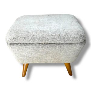 May include: A square ottoman upholstered in a textured, light gray woven fabric. The piece features a slightly tapered, boxy seat cushion and is supported by four short, angled wooden legs with a smooth, medium-brown polished finish.