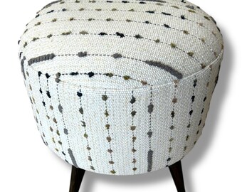 May include: A round ottoman featuring a cream-colored woven fabric with vertical stripes of small, raised tufted dots in shades of gray, brown, and black. The piece is supported by three dark brown wooden legs that taper toward the floor.