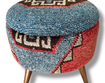 May include: A round ottoman featuring a textured, woven fabric upholstery with a geometric pattern in shades of blue, red, and dark brown. The seat is supported by three short, tapered wooden legs with a smooth, natural brown finish.