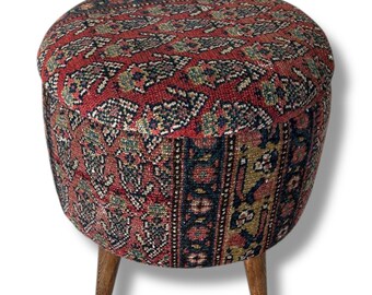 May include: A round ottoman upholstered in a woven fabric featuring a red, blue, and gold patterned design. The seat is cylindrical with a flat top and is supported by three short, tapered wooden legs with a natural brown finish.