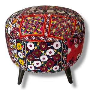 May include: A round ottoman featuring a patchwork fabric cover with vibrant red, yellow, and blue embroidered floral and geometric patterns. The seat is supported by three dark brown tapered wooden legs, providing a stable base for the stool.