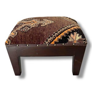 May include: A square ottoman featuring a dark brown, textured woven top with geometric patterns in orange, black, and cream. The base is constructed from dark stained wood with four sturdy legs and a row of silver-toned nailhead trim accents.