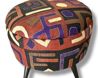 May include: A round ottoman featuring a textured fabric upholstery with a geometric pattern in shades of black, rust orange, cream, and purple. The seat is supported by three short, tapered, dark brown wooden legs, providing a stable base.