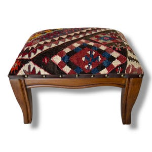 May include: A low footstool with a rectangular top upholstered in a woven fabric featuring a multi-colored geometric pattern in red, blue, black, and cream. The base is made of polished brown wood with four curved legs and metal nailhead trim.