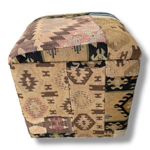 May include: A square ottoman upholstered in a patchwork of woven fabric featuring various geometric patterns in shades of tan, brown, black, and muted pink. The textile surface displays a repeating arrangement of diamonds, stripes, and angular motifs.