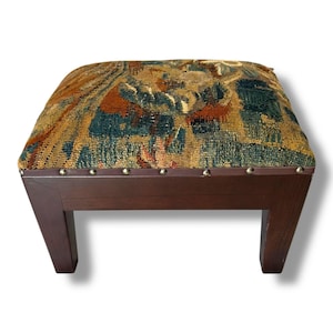 May include: A square footstool featuring a dark brown wooden base with four tapered legs. The top is upholstered in a multi-colored woven tapestry fabric displaying intricate patterns in shades of blue, rust, gold, and tan, secured with silver nailhead trim.