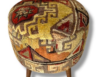 May include: A round ottoman upholstered in a textured, multi-colored woven fabric featuring geometric patterns in shades of red, yellow, orange, brown, and cream. The piece is supported by three short, tapered wooden legs with a visible grain finish.