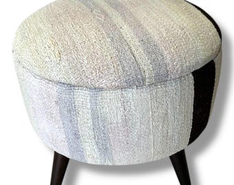 May include: A round ottoman featuring a textured, woven fabric upholstery in shades of cream, light gray, and soft blue, accented by a vertical dark brown stripe. The piece is supported by three tapered, dark brown wooden legs.
