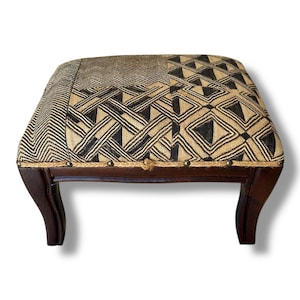 May include: A square footstool with dark brown wooden legs and a textured fabric top featuring a black and beige geometric pattern. The upholstery is secured with a row of metallic nailhead trim along the edge of the wooden frame base.