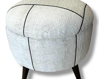 May include: A round ottoman upholstered in a textured off-white fabric with a visible black grid pattern. The piece features a flat circular top and a cylindrical body supported by three dark brown tapered wooden legs that extend outward.