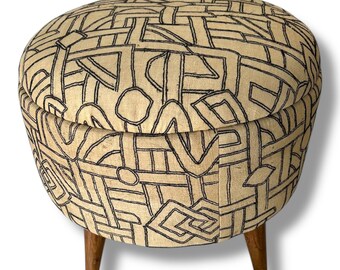 May include: A round ottoman featuring a beige fabric upholstery with a black embroidered geometric pattern of intersecting lines and shapes. The piece is supported by three short, tapered wooden legs in a medium brown finish.
