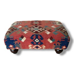 May include: A rectangular footstool featuring a woven fabric top with a geometric pattern in shades of red, blue, white, and brown. The piece is supported by four dark brown, rounded wooden feet, providing a stable base for the upholstered top.