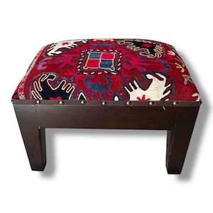 May include: A square footstool with a dark brown wooden frame and four legs. The top is upholstered in a red fabric featuring a woven pattern of black and white animal-like shapes and a central blue and red square motif, accented by metal studs.
