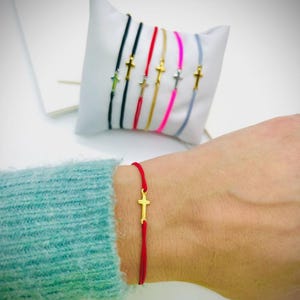 以下が含まれることがあります： 赤い紐のブレスレットに小さな金の十字架のチャームが付いている。ブレスレットは人の手首につけられている。