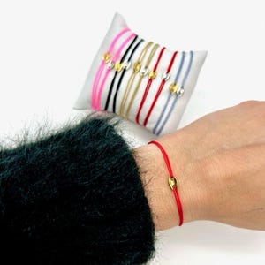 May include: A red string bracelet with a gold charm. The bracelet is on a person's wrist. There are other bracelets in different colors on a white display stand.