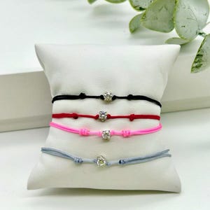 Puede incluir: Cuatro pulseras con cordones de diferentes colores y un pequeño dije plateado con una piedra transparente en el centro. Las pulseras se muestran en una almohada blanca.