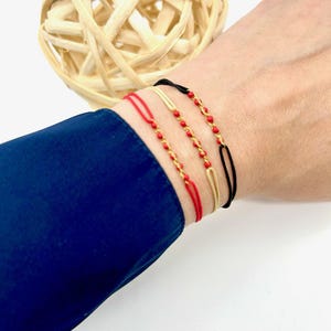 May include: Three bracelets on a wrist. The bracelets are red string with gold beads, a gold chain, and a black string with a gold bar.