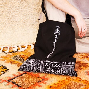 May include: Black tote bag with white embroidered geometric patterns. The bag has a long strap and is made of canvas.