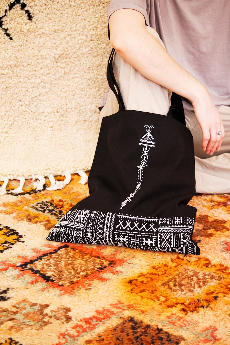May include: Black tote bag with white embroidered geometric patterns. The bag has a long strap and is made of canvas.