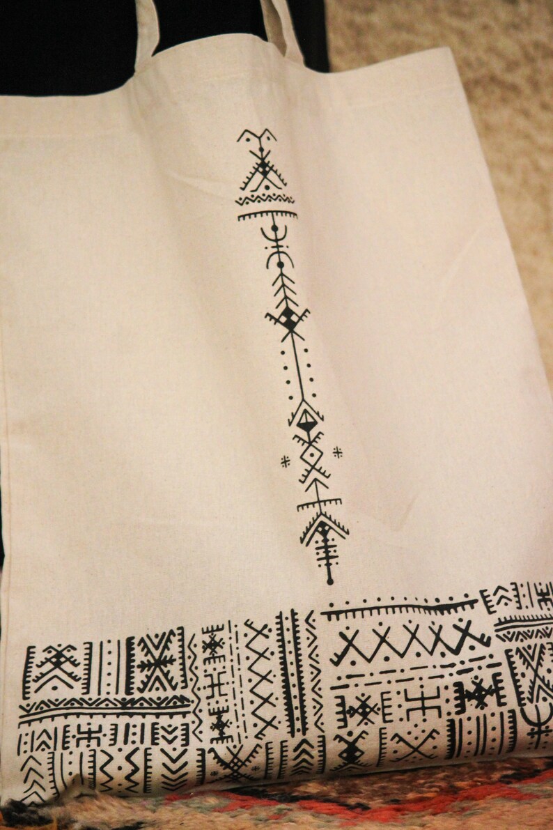 May include: A white canvas tote bag with black geometric designs. The design features a vertical pattern of triangles, diamonds, and dots. The bottom of the bag has a horizontal pattern of geometric shapes.