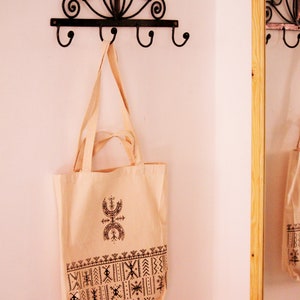 May include: A white canvas tote bag with black geometric patterns and a symbol printed on the front. The bag is hanging on a black metal hook with a decorative design.
