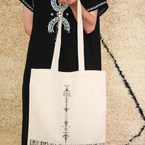 May include: A white canvas tote bag with black geometric designs. The bag has a long strap and is held by a person wearing a black dress with white embroidery.