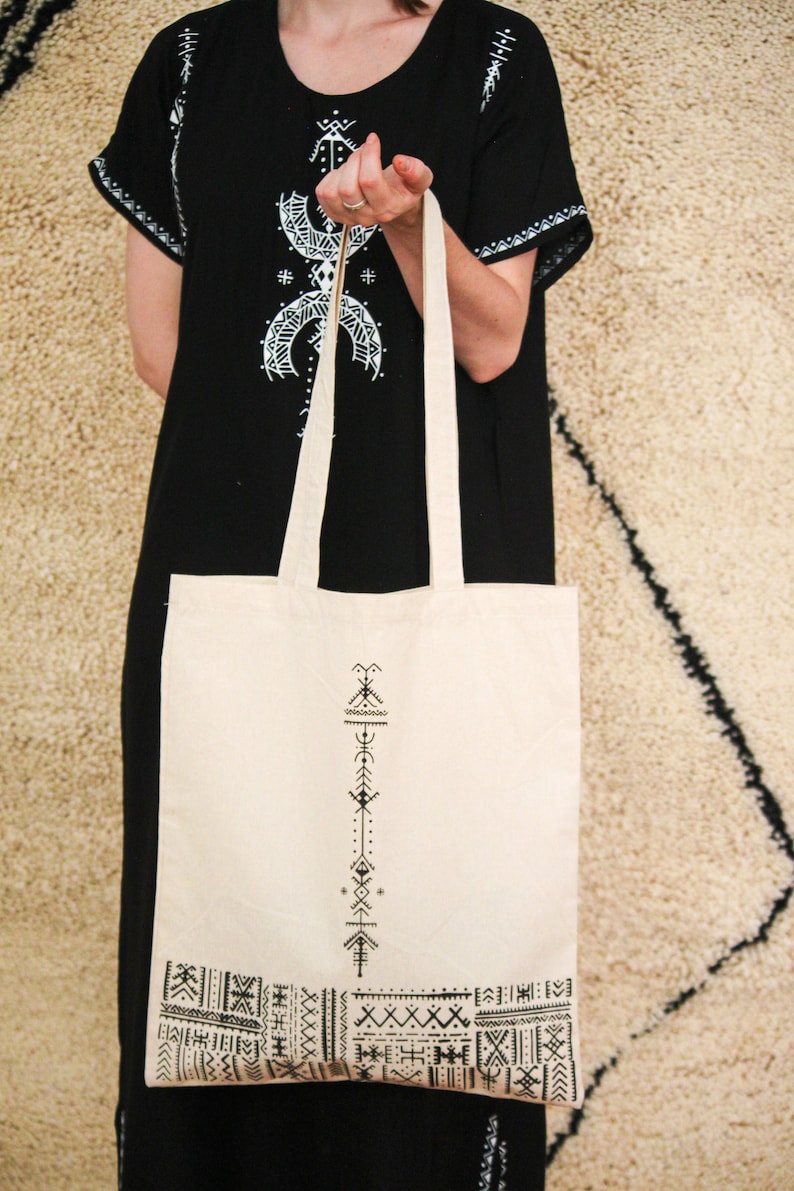 May include: A white canvas tote bag with black geometric designs. The bag has a long strap and is held by a person wearing a black dress with white embroidery.